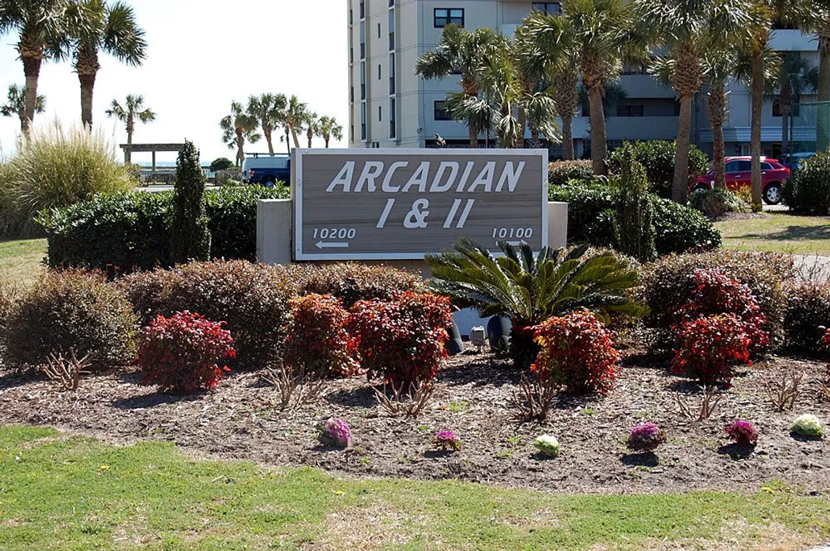 A welcoming view of the Arcadian I & II sign situated among lush landscaped gardens and palm trees, with surrounding resort buildings visible. The setting feels inviting and tropical, emphasizing the location’s appeal.