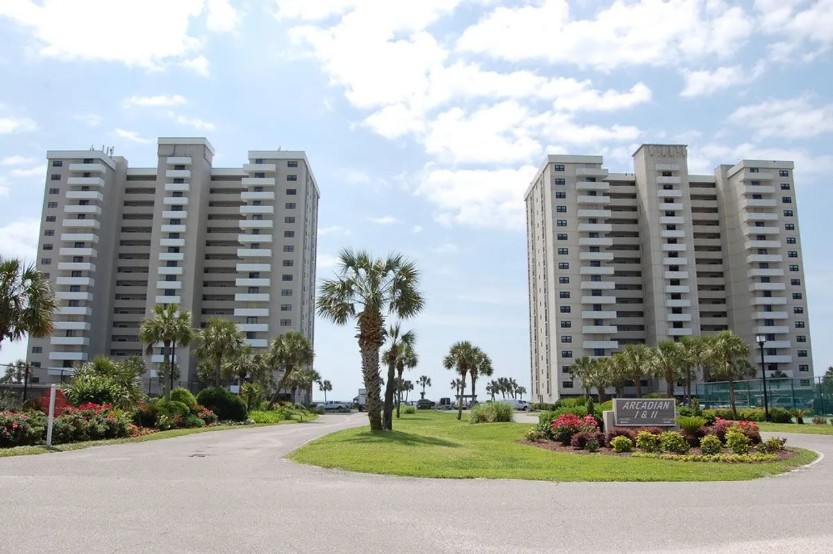 An inviting exterior view of two high-rise buildings surrounded by landscaped gardens and palm trees. The scene includes a welcoming entrance and hints at on-site amenities like outdoor social spaces, suggesting comfort, privacy, and relaxation.