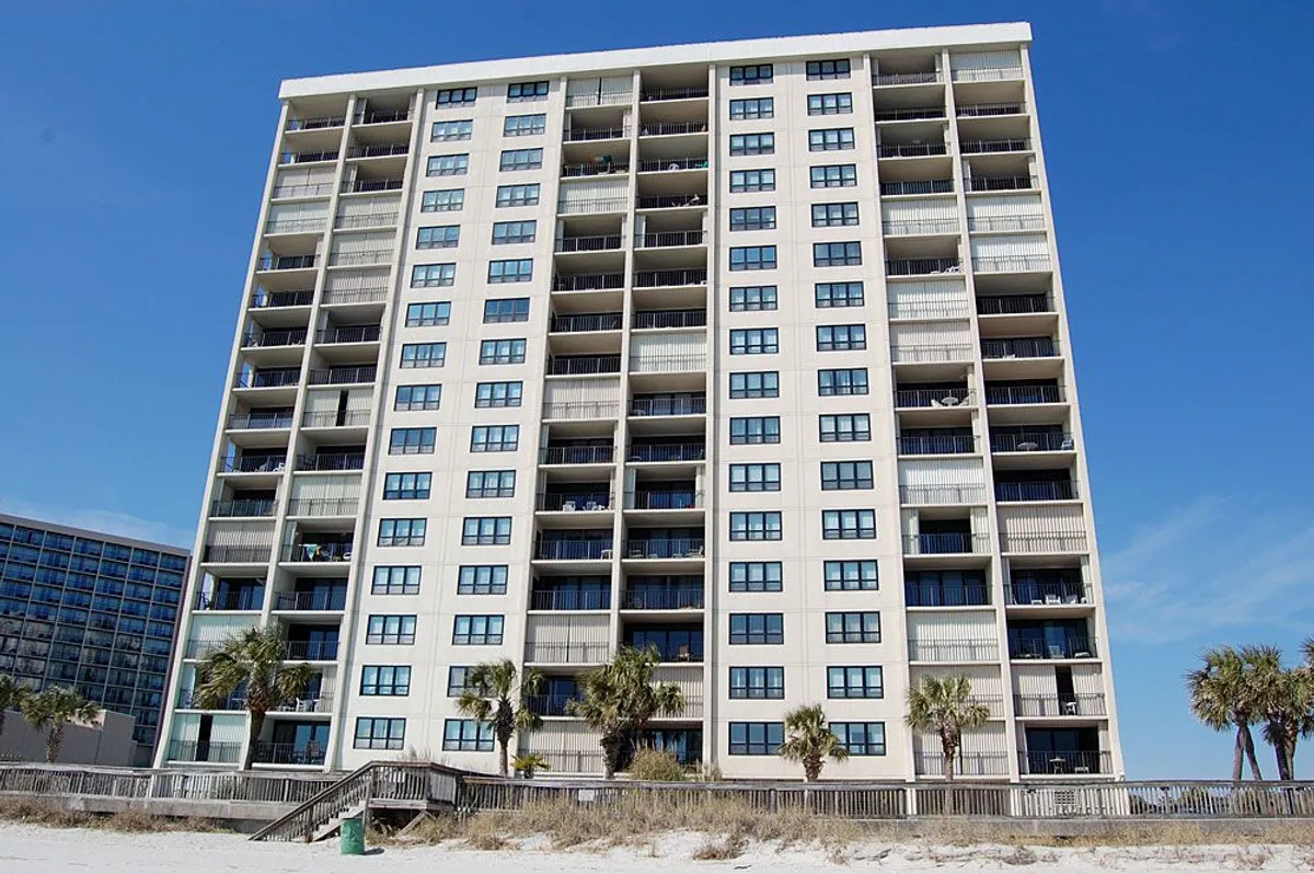 A tall condominium building with rows of balconies overlooking the beachfront, framed by swaying palm trees. The setting conveys a relaxed, coastal resort atmosphere, showcasing the property’s proximity to the ocean and inviting outdoor lifestyle.