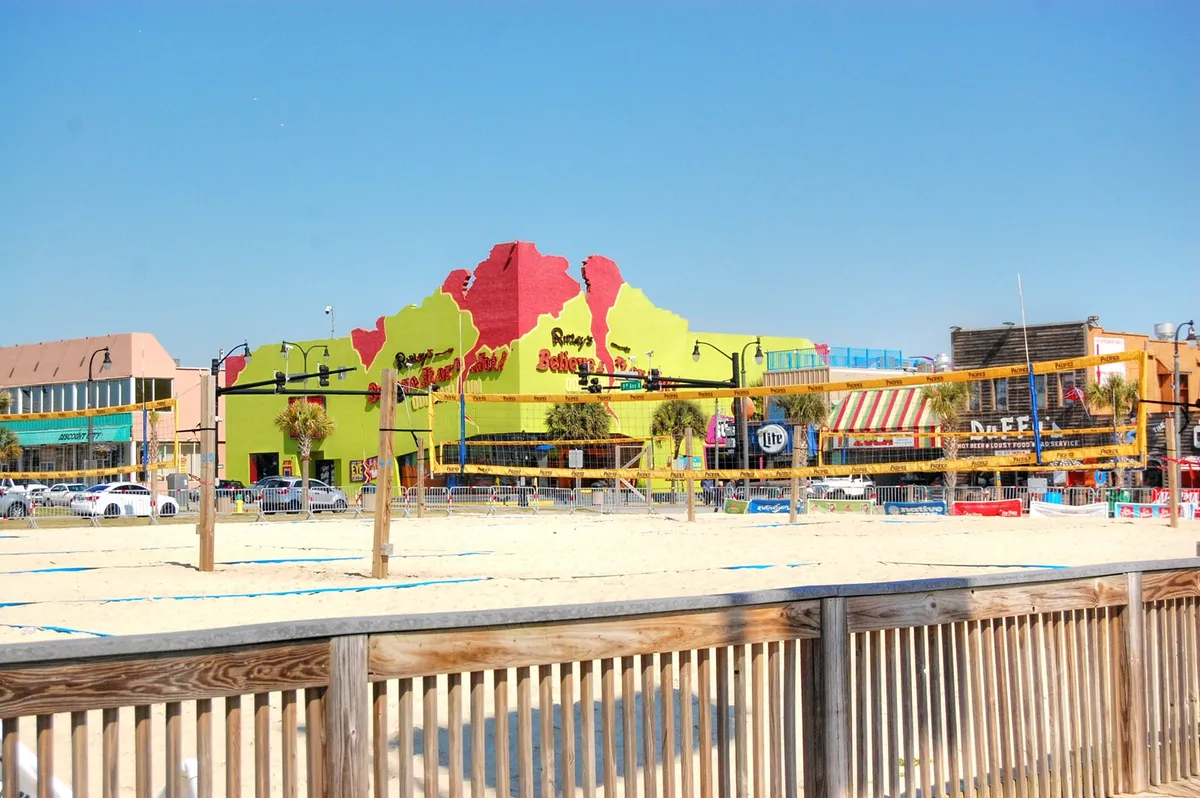 A colorful exterior shot featuring a building adjacent to beach volleyball courts, framed by palm trees and inviting signage. The area buzzes with activity, offering opportunities for dining, shopping, and socializing—ideal for groups seeking fun experiences.