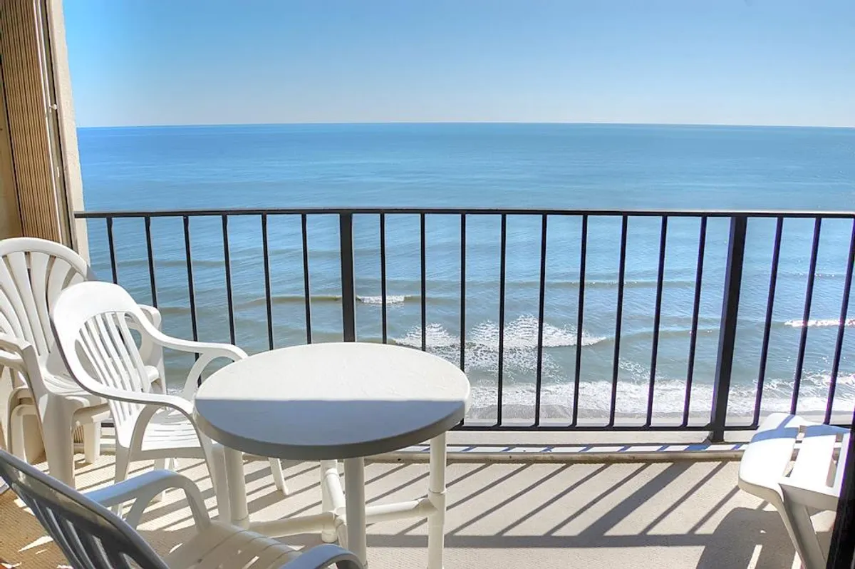 A serene view from the balcony with a table and chairs overlooking the ocean. The scene is set for enjoying a meal or coffee while taking in the tranquil sunrise or the gentle sound of the waves. The furniture arrangement invites dining or lingering conversations outdoors.