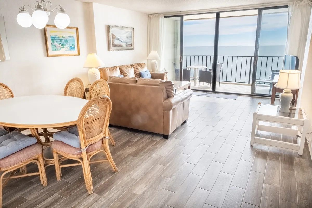 A bright, inviting living and dining area featuring a comfortable couch, large window framing a stunning ocean view, a dining table ready for gatherings, and decorative paintings that add warmth to the space. Natural light pours in, accentuating the sense of openness and connection to the coastal landscape.