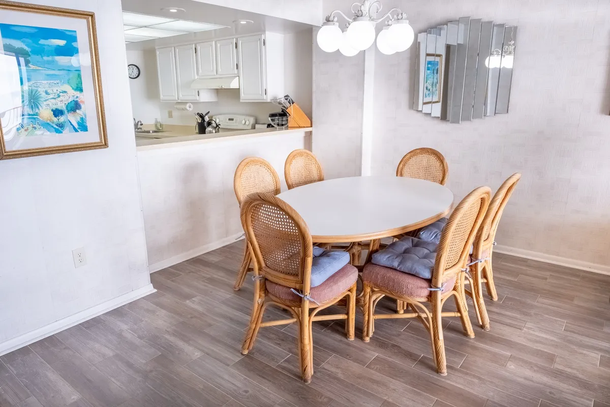 A stylish dining area with a round table surrounded by woven chairs, a decorative mirror, kitchen pass-through, and a warm chandelier overhead. The setting is perfect for intimate meals, board games, or lively conversation with friends and family.