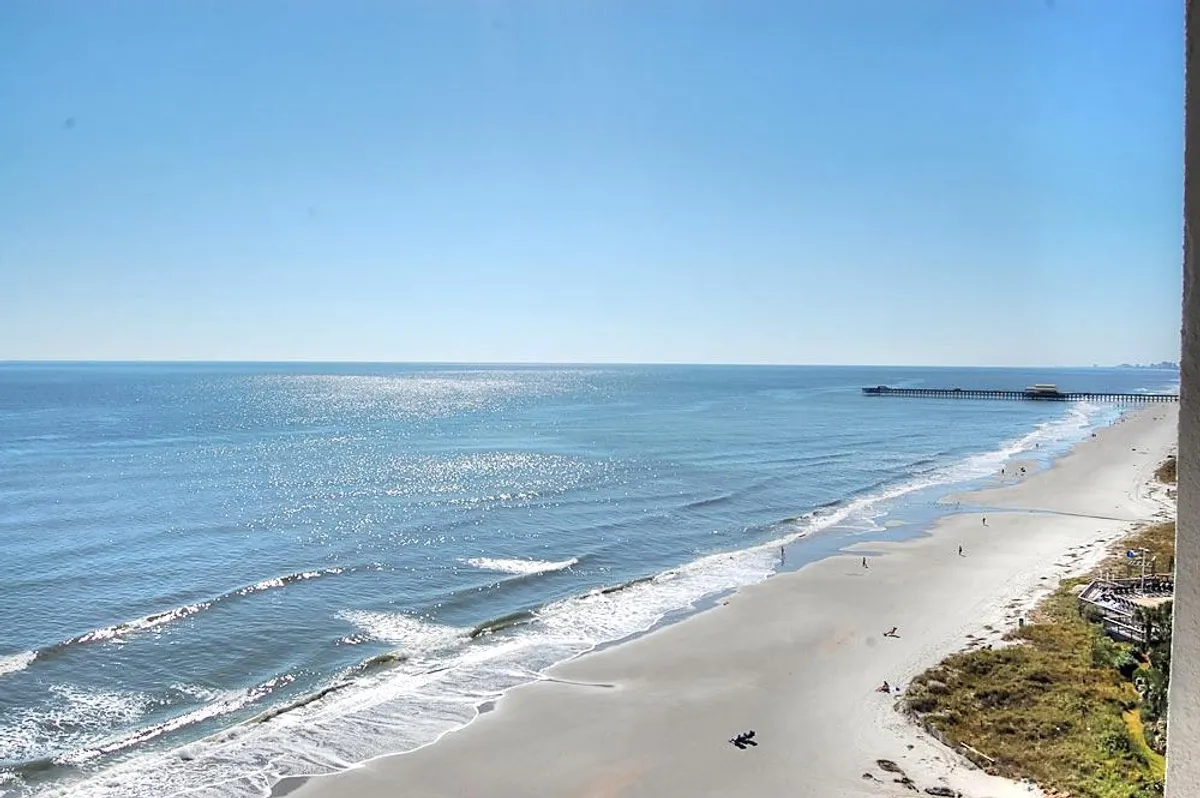 A stunning view of ocean waves meeting a sandy beach, with a pier stretching into the distance and sunlight sparkling on the water. The serene, open space invites beach walks and outdoor relaxation.