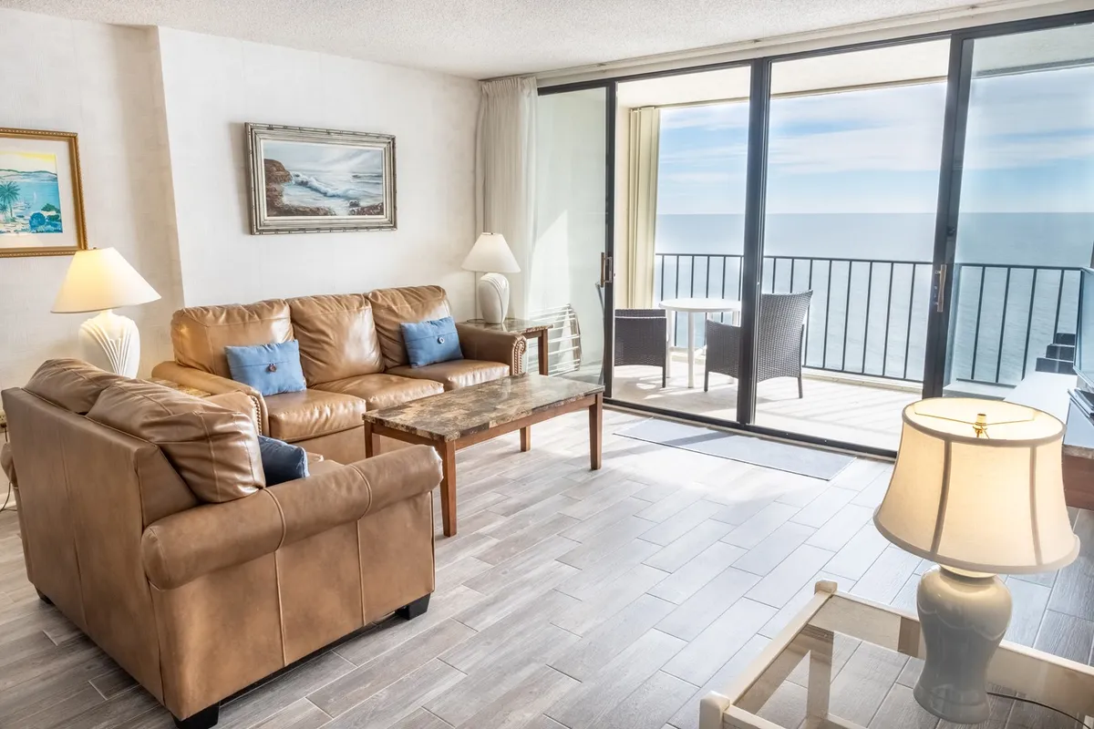 The image showcases a stylish and comfortable living room featuring a leather sofa and coffee table, warmly lit by lamps. Large glass doors open out to a private balcony, offering breathtaking ocean views and inviting natural light that fills the space, hinting at serene evenings and stunning sunsets.