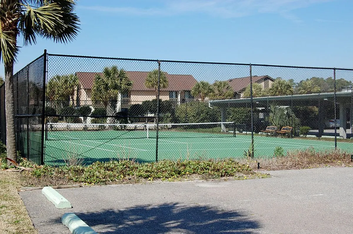 An outdoor tennis court bordered by lush palm trees, with residential buildings visible in the background. The sunlit court is ready for friendly matches or solo practice, providing an active leisure option within the resort grounds.