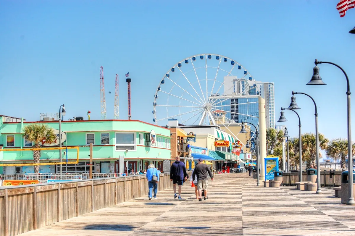 A vibrant boardwalk scene featuring a towering ferris wheel, colorful buildings, palm trees, and families enjoying amusement rides and dining. The atmosphere is lively, playful, and distinctly Myrtle Beach.