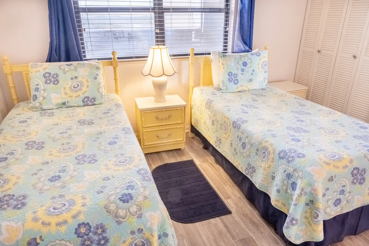 A cheerful bedroom with two twin beds, a lamp, a window with curtains, and a dresser. The space exudes warmth and comfort, ideal for kids or friends sharing the room.