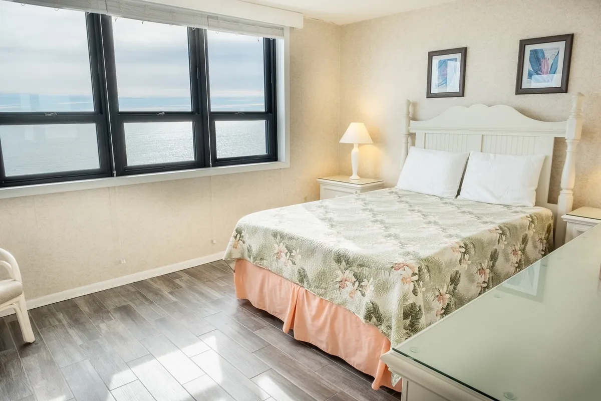 A tranquil bedroom featuring a bed with floral bedding, large windows offering an ocean view, a nightstand with a lamp, and framed artwork on the walls. Soft natural light fills the space, enhancing the peaceful ambiance.